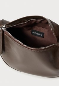 Pungă maro din piele cu un fermoar argintiu; exterior neted și interior moale din material textil; etichetă interioară vizibilă cu numele brandului.