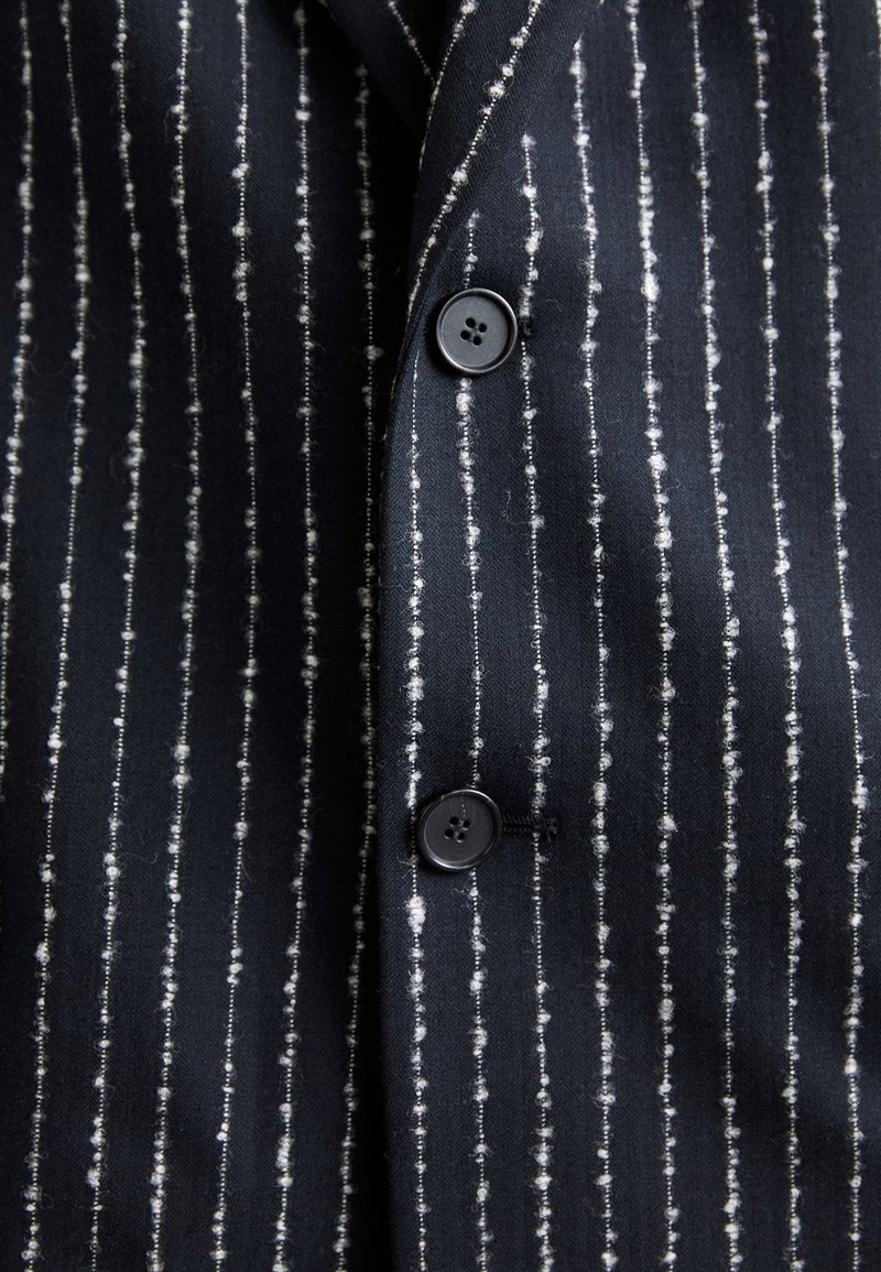 Granatowa koszula w białe pionowe paski, wykonana z teksturowanej tkaniny. Posiada czarne guziki oraz klasyczny kołnierzyk.