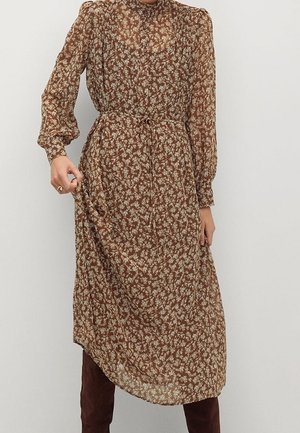 Hnědé midi šaty s dlouhým rukávem a bílým květinovým potiskem, se staženým pasem a zapnutým límečkem, nošené s tmavě hnědými botami.