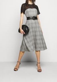 Robe à carreaux gris avec un haut transparent noir, une taille ceinturée, une jupe fluide, et accompagnée d'une pochette noire et de talons à lanières.