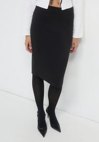 Frau trägt einen schwarzen asymmetrischen Bleistiftrock, schwarze Strumpfhosen, einen weißen strukturierten, kurzen Pullover und schwarze spitze High Heels mit Schnürdetails.