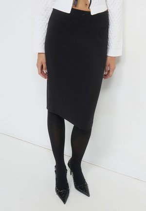 Frau trägt einen schwarzen asymmetrischen Bleistiftrock, schwarze Strumpfhosen, einen weißen strukturierten, kurzen Pullover und schwarze spitze High Heels mit Schnürdetails.