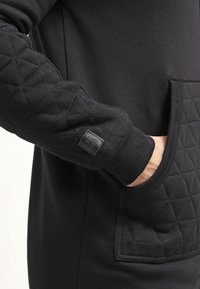Felpa nera con texture trapuntata sulle maniche e tasca anteriore, con un'etichetta cucita e polsini a costine.