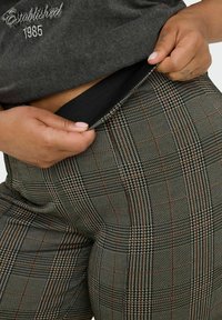 Pantalon à carreaux avec une ceinture élastique noire, principalement dans des nuances de gris avec des accents orange et blanc, présentant un tissu texturé.