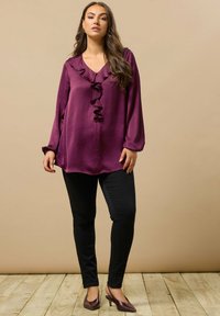 Bordeauxfarbene Bluse mit Rüschenkragen, langen Ärmeln und weicher Textur, kombiniert mit schwarzen schmalen Hosen und spitzen Pumps.