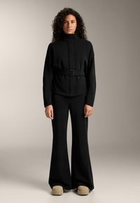 Crna jakna dugih rukava s pojasom i visokim ovratnikom, u kombinaciji s crnim hlačama na zvono i bež platformama. Glatka tekstura, minimalistički dizajn.