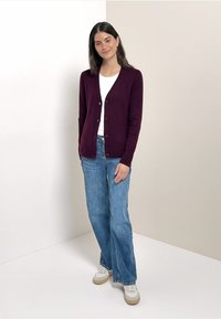 Burgundivärinen villatakki V-kaula-aukolla ja nappisulkimella, käytetty valkoisen t-paidan päällä, yhdistettynä vaaleansinisiin leveälahkeisiin farkkuihin ja valkoisiin tennareihin.