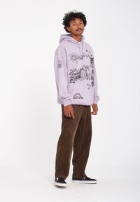 Sweat à capuche lavande avec des illustrations graphiques noires, associé à un pantalon en velours côtelé marron et des baskets noires. Le sweat à capuche possède une poche kangourou.