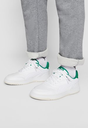 Hvide og grønne Puma sneakers båret med hvide Puma sokker og opkørte grå joggingbukser på en neutral baggrund.