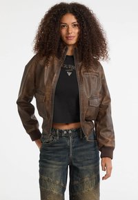 Bruine leren bomberjack met ribgebreide manchetten, ritssluiting aan de voorkant en twee voorzakken. Draagt over een zwart cropped top en gecombineerd met gestructureerde denim jeans.