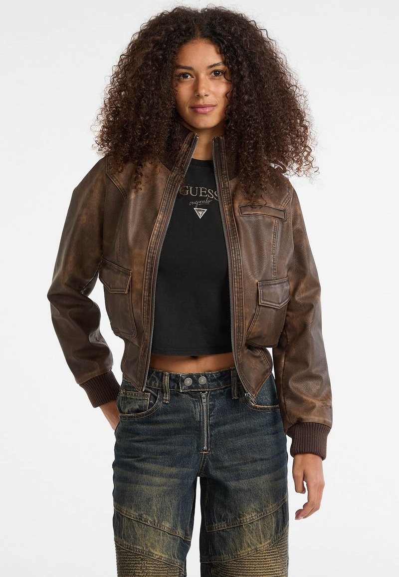 Bruine leren bomberjack met ribgebreide manchetten, ritssluiting aan de voorkant en twee voorzakken. Draagt over een zwart cropped top en gecombineerd met gestructureerde denim jeans.