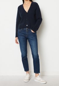 Marineblauer gerippter Strickcardigan mit V-Ausschnitt und vorderen Knöpfen, kombiniert mit dunkelblauen knöchellangen Jeans und weißen Sneakern.