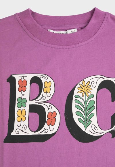 Lilla T-shirt med store dekorative bogstaver "B" og "C", der indeholder farverige blomster- og bladdesigns inden i bogstaverne.