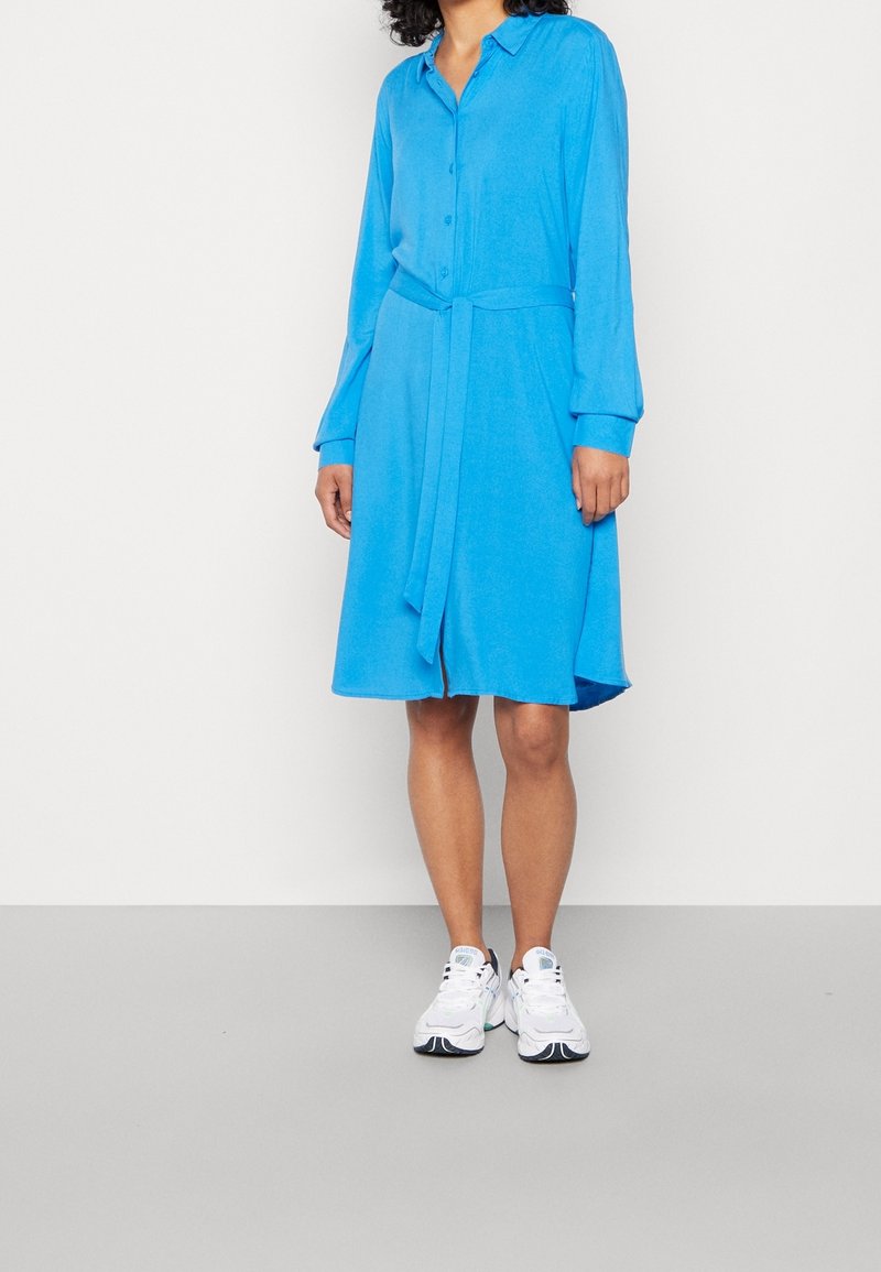 Femme portant une robe chemise bleu vif à manches longues avec une ceinture à la taille et des baskets blanches, debout devant un fond uni.