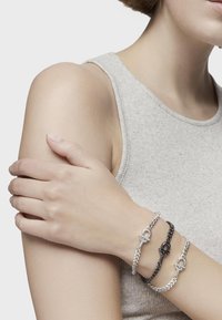 Pulseras de cadena plateadas y negras con cierres de palanca, llevadas en un brazo desnudo, en capas sobre una blusa de textura gris clara.