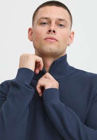 Temno modra zip-up pulover iz strukturirane tkanine, z visokim ovratnikom in rebrastimi manšetami, z gladkim zadrgo podrobnostjo na vratu.