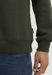 Sudadera verde oscura con puños y dobladillo de canalé, mostrando una tela texturizada. Mano descansando sobre unos vaqueros con detalles de costura visibles.