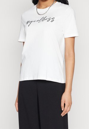 Personne portant un t-shirt blanc à manches courtes avec « #girlboss » imprimé en cursive noire et un pantalon noir, accessoirisée d'un collier chaîne en argent.