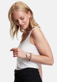 Guldtonet ur med en rund urskive og leddet kædearmbånd; parret med en kraftig kædehalssmykke; model iført en hvid tanktop.