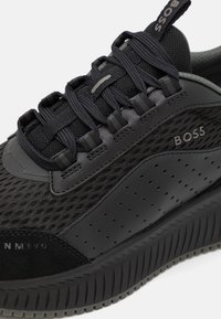 Chaussure de sport noire alliant mesh et cuir, avec une semelle extérieure texturée et un marquage "BOSS" bien visible.