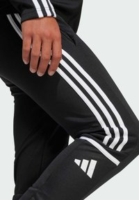 Czarne spodnie sportowe z białymi paskami po bokach. Charakteryzują się gładką fakturą i logo na dolnej części nogawki. Zbliżenie na materiał i dopasowanie.