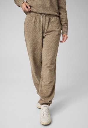 Person iført beige leopardmønstrede joggingbukser og matchende top med hvide sneakers, der går mod en ensfarvet baggrund.