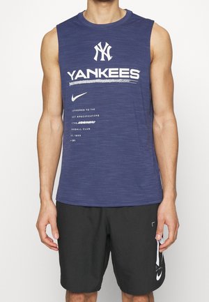 Homme portant un maillot sans manches bleu marine des Yankees et un short noir Nike, debout devant un fond uni.