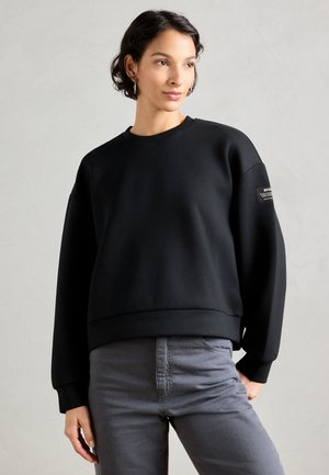Sudadera negra con cuello redondo, corte holgado y mangas largas con puños. Presenta un pequeño parche de logo en la manga izquierda.