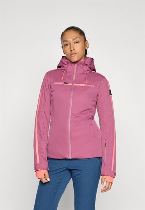 Frau in einer lila Kapuzenjacke mit pinken Streifen und blauen Hosen, die vor einem einfarbigen hellen Hintergrund steht.