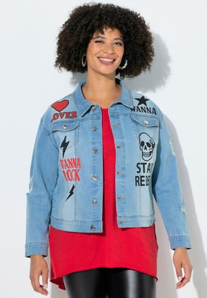 Smilende person med krøllet hår iført en lyseblå denimjakke med røde og sorte rock-tema patches over en rød skjorte og sorte leggings.