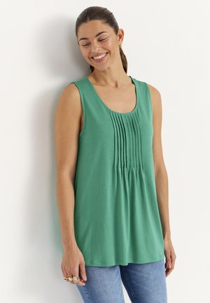 Kvinde smiler med lukkede øjne, iført ærmeløs grøn top med lodrette plissé og blå jeans, står foran en hvid væg.