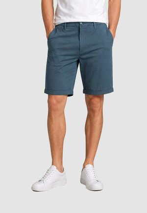 Uomo che indossa pantaloncini blu con orlo arrotolato, sneaker bianche e una camicia bianca, in piedi con le mani nelle tasche su uno sfondo chiaro e semplice.