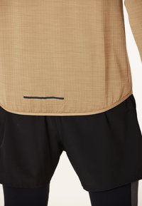 Tan langærmet atletisk top med struktureret stof og sort kantlinje i kanten, parret med sorte shorts lavet af glat materiale.