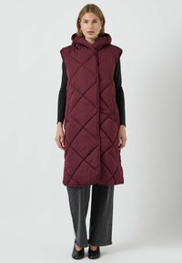 Burgundy kviltad lång väst med huva, med diagonala sömmönster och öppen front. Bärs över en svart långärmad topp och grå byxor.