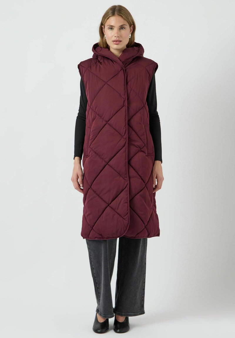 Burgundy kviltad lång väst med huva, med diagonala sömmönster och öppen front. Bärs över en svart långärmad topp och grå byxor.