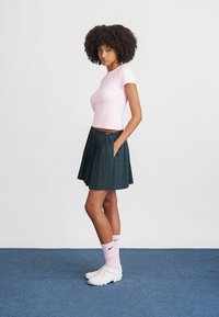 Gaiši rozā īsām piedurknēm krekliņš apvienots ar tumši zaļu plisēto svārku. Modele valkā rozā zeķes ar melnu logo un baltas kurpes.