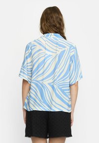 Camicia a maniche corte in azzurro chiaro e crema con righe ondulate; vestibilità comoda, tessuto morbido e dettaglio della spalla sul retro. Abbinata a pantaloni corti neri.