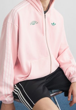Person iført en lyserød Adidas-hættetrøje med grønne logoer og sorte shorts med hvide striber, der sidder på en stol.