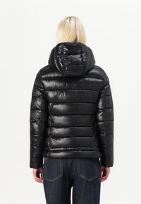Svart dunjakke med glanset overflate, horisontalt quiltet design, hette og lange ermer, paret med mørke jeans, sett fra ryggen.