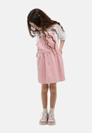 Jeune fille aux longs cheveux bouclés, portant une robe tablier à rayures rose et blanches, une chemise blanche et des baskets roses, regardant vers le bas.