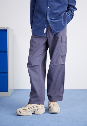 Person wearing loose gray cargo pants, navy blue buttoned shirt, and beige chunky closed-toe sandals standing on blue carpet.