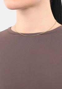 Un collar de cadena de oro reposa sobre un escote redondeado de una blusa ajustada y suave en tono malva. La blusa tiene un ligero brillo y un diseño minimalista.