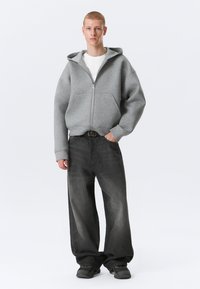 Junge Person, die einen grauen Kapuzenpullover mit Reißverschluss, ein weißes Hemd, übergroße schwarze Jeans und schwarze Turnschuhe trägt und mit einer Hand in der Tasche steht.