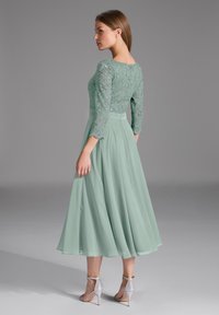 Robe vert menthe avec bodice en dentelle, manches trois-quarts et jupe plissée fluide. Des accents métalliques sur les sandales ajoutent du détail.