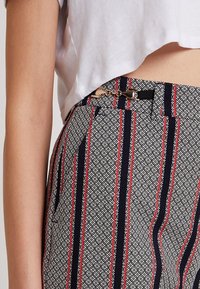 Kvinna som bär mönstrade byxor med svarta, vita och röda vertikala ränder och en guldkrok, som kombineras med en kortärmad vit topp som visar midjan.