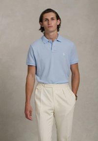 Νεαρός άνδρας που φοράει ανοιχτό μπλε πόλο και κρεμ παντελόνι με πτυχώσεις, στέκεται μπροστά σε απλή μπεζ βαφή.