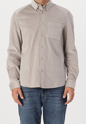 Camisa de pana de color marrón claro con cuello abotonado, bolsillo frontal y textura acanalada. Mangas largas enrolladas en los puños, combinada con vaqueros azules.