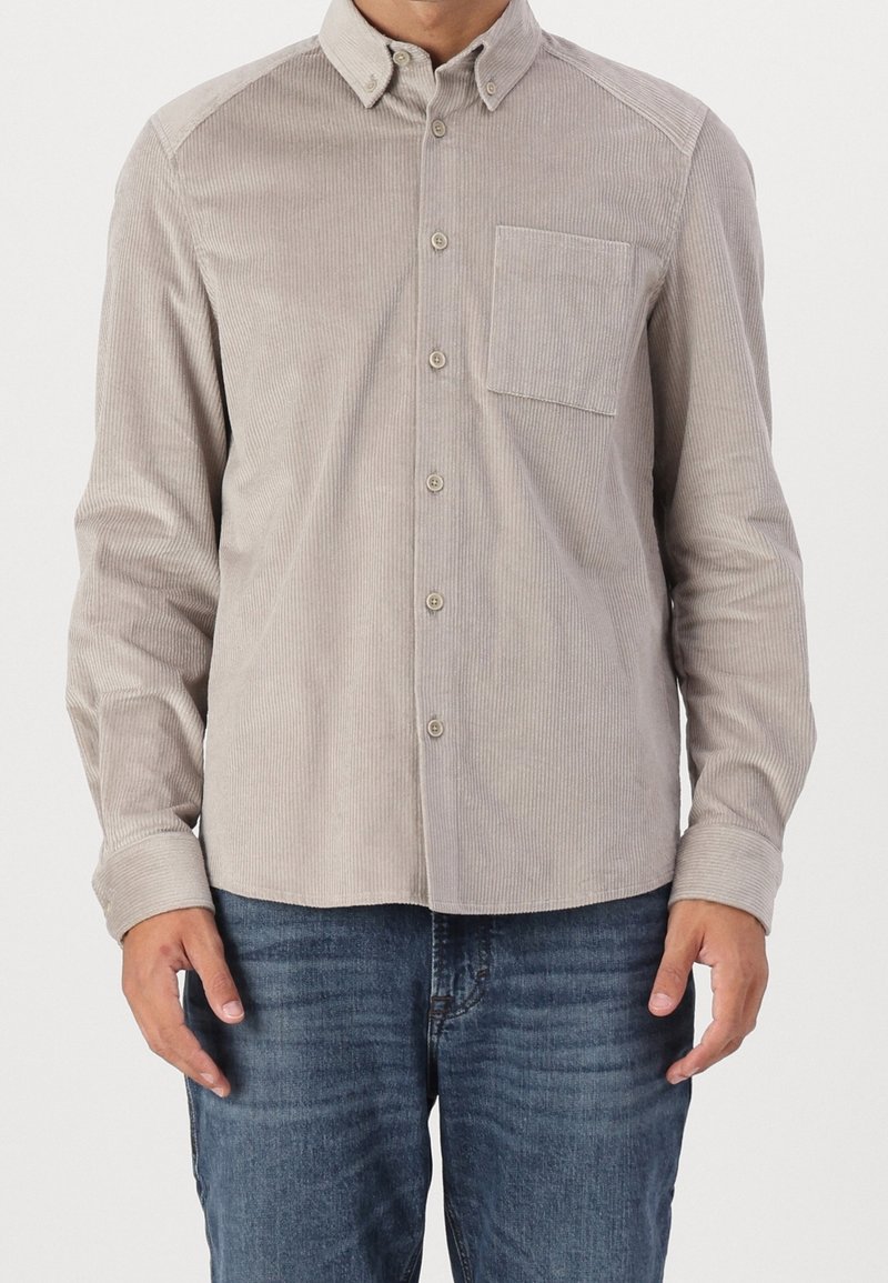 Camisa de pana de color marrón claro con cuello abotonado, bolsillo frontal y textura acanalada. Mangas largas enrolladas en los puños, combinada con vaqueros azules.