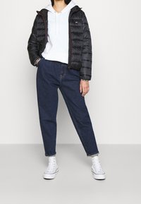 Chaqueta de plumas negra, textura acolchada, cierre con cremallera, combinada con una sudadera gris claro y pantalones de mezclilla azul de cintura alta. Zapatillas blancas.