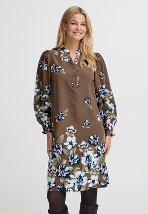 Femme en robe marron avec de grands motifs floraux blancs, bleus et verts, manches longues bouffantes, collants foncés et bottes foncées, souriante à la caméra.